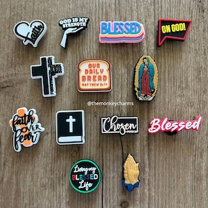 May include: A selection of shoe charms with religious motifs. The charms feature designs such as crosses, hearts, and phrases like "God is My Strength" and "Living My Blessed Life." The charms are in various colours and styles.