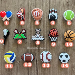 K&ouml;nnte beinhalten: Eine Sammlung von Sport-Themen-Anh&auml;ngern, darunter Tischtennisschl&auml;ger, ein Baseball und Schl&auml;ger, ein Baseballhandschuh, Bowlingkegel, ein Tennisball, ein Lacrosseschl&auml;ger, eine Feldhockeyszene, Hockeyschl&auml;ger und ein Puck, ein Basketball in einem Korb, ein Basketball, ein Volleyball, ein Football, ein Baseball und ein Fu&szlig;ball.