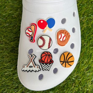 K&ouml;nnte beinhalten: Wei&szlig;er Clog-Schuh mit Sport-Themen-Anh&auml;ngern. Zu den Anh&auml;ngern geh&ouml;ren ein Baseball mit Schl&auml;ger, Tischtennisschl&auml;ger, ein Baseball, ein Baseballhandschuh, Hockeyschl&auml;ger und Puck, ein Basketball mit Korb und ein Basketball.
