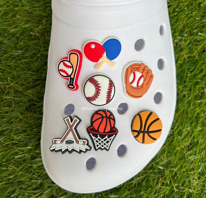 K&ouml;nnte beinhalten: Wei&szlig;er Clog-Schuh mit Sport-Themen-Anh&auml;ngern. Zu den Anh&auml;ngern geh&ouml;ren ein Baseball mit Schl&auml;ger, Tischtennisschl&auml;ger, ein Baseball, ein Baseballhandschuh, Hockeyschl&auml;ger und Puck, ein Basketball mit Korb und ein Basketball.