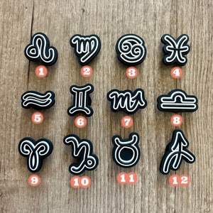May include: A collection of twelve black and white zodiac symbol charms. Each charm features a unique astrological sign design with a white outline. The charms are arranged on a light brown wooden surface.