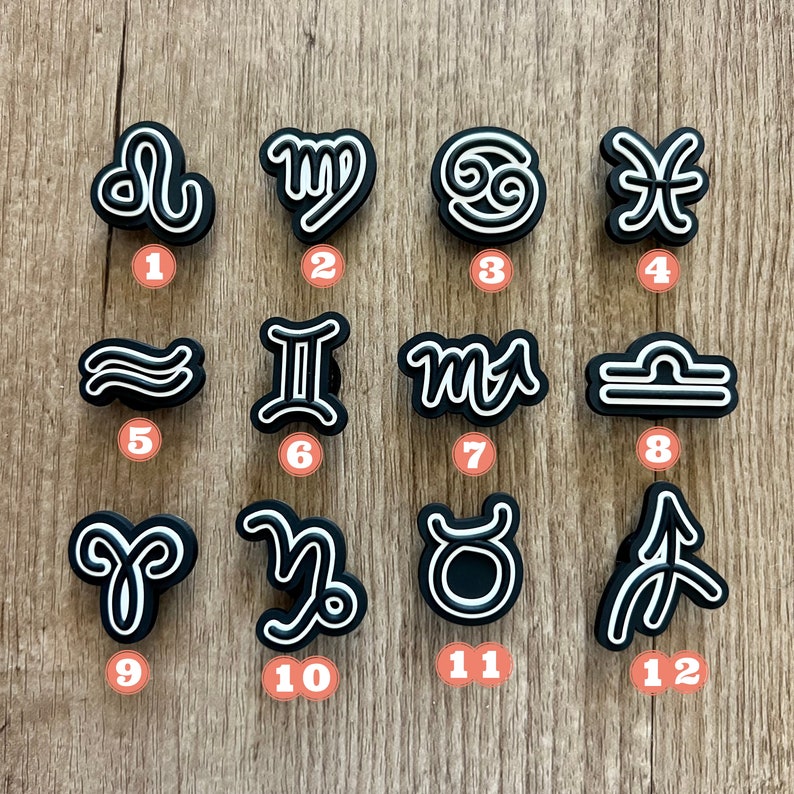 May include: A collection of twelve black and white zodiac symbol charms. Each charm features a unique astrological sign design with a white outline. The charms are arranged on a light brown wooden surface.
