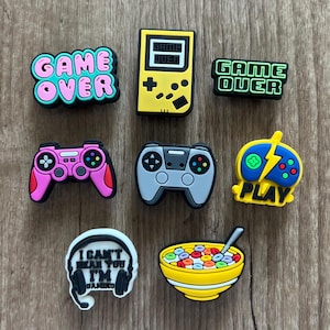 May include: A collection of colorful gaming-themed charms. Includes charms shaped like game controllers, a handheld game console, and the words "GAME OVER" and "PLAY". Also includes a charm with the text "I CAN'T HEAR YOU I'M GAMING" and a bowl of cereal.