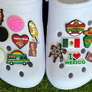 May include: White clogs decorated with colorful charms. Charms include a taco truck, a heart with a taco, a Mexican flag, a pinata, and the words "I Love Mexico". Other charms include a corn on the cob and a concha.