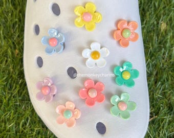 May include: White shoe with colorful flower charms. The charms are in various pastel colors, including pink, yellow, blue, and green. Each flower has a contrasting center. The shoe is on a green grassy surface.