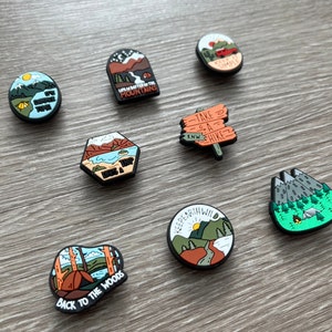 May include: A collection of camping-themed shoe charms with various designs. The charms feature images of mountains, tents, and outdoor scenes, with phrases like "Life's a Trip" and "Back to the Woods."
