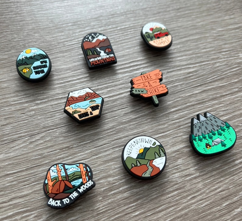May include: A collection of camping-themed shoe charms with various designs. The charms feature images of mountains, tents, and outdoor scenes, with phrases like "Life's a Trip" and "Back to the Woods."