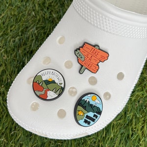 May include: White clog shoe with three decorative charms. One charm says "KEEP EARTH WILD" with a mountain scene. Another charm says "TAKE A HIKE" on a wooden sign. The third charm says "IT'S SUMMER TIME" with a mountain and tent scene.