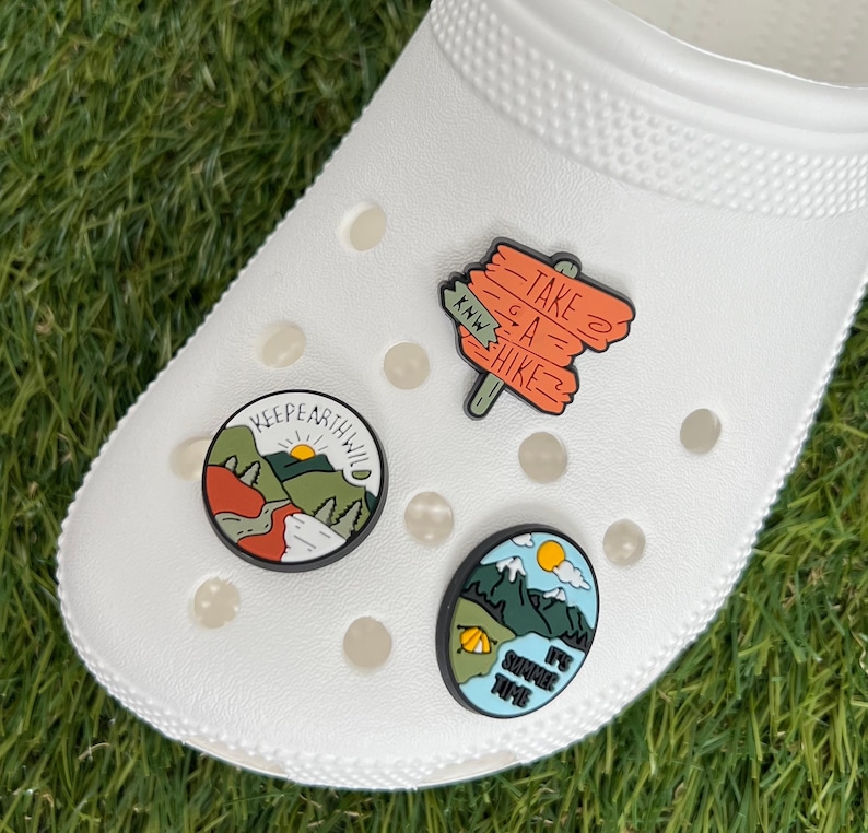 May include: White clog shoe with three decorative charms. One charm says "KEEP EARTH WILD" with a mountain scene. Another charm says "TAKE A HIKE" on a wooden sign. The third charm says "IT'S SUMMER TIME" with a mountain and tent scene.