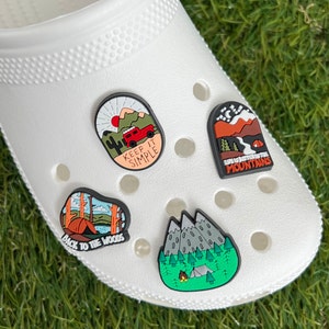 May include: White clog shoe with four camping-themed charms. Charms include a red jeep with the text "KEEP IT SIMPLE", a mountain scene with the text "LIFE IS BETTER IN THE MOUNTAINS", a tent with the text "BACK TO THE WOODS", and a mountain scene with a campfire.