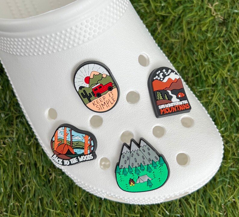 May include: White clog shoe with four camping-themed charms. Charms include a red jeep with the text "KEEP IT SIMPLE", a mountain scene with the text "LIFE IS BETTER IN THE MOUNTAINS", a tent with the text "BACK TO THE WOODS", and a mountain scene with a campfire.
