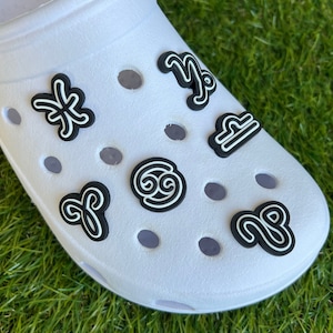 May include: White clog shoe adorned with black and white zodiac sign charms. The charms include symbols for Pisces, Capricorn, Libra, Cancer, Aries, and Sagittarius. The shoe is on a green grassy surface.