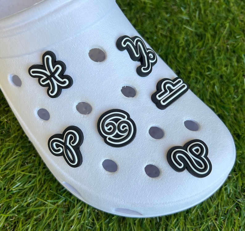 May include: White clog shoe adorned with black and white zodiac sign charms. The charms include symbols for Pisces, Capricorn, Libra, Cancer, Aries, and Sagittarius. The shoe is on a green grassy surface.