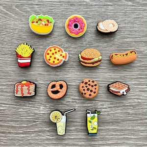 May include: A collection of colorful food-themed charms, including a salad, donut, bagel, french fries, pizza, burger, hot dog, pancakes, pretzel, cookie, sandwich, and two beverages. The charms are made of a glossy material and are arranged on a wooden surface.