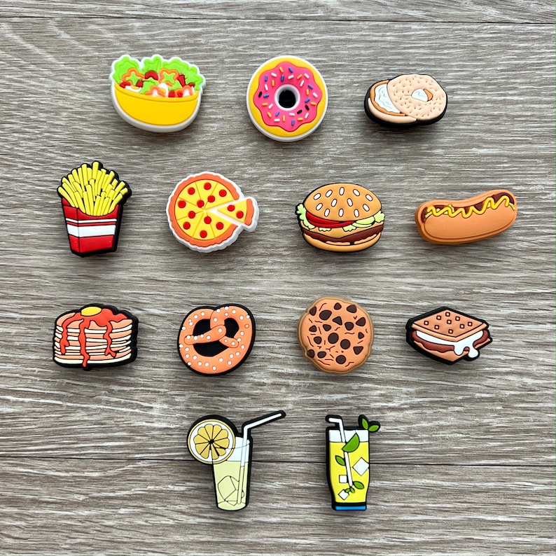 May include: A collection of colorful food-themed charms, including a salad, donut, bagel, french fries, pizza, burger, hot dog, pancakes, pretzel, cookie, sandwich, and two beverages. The charms are made of a glossy material and are arranged on a wooden surface.