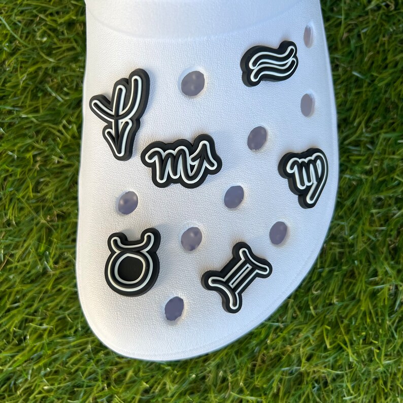 May include: White clog shoe adorned with black and white zodiac sign charms. The charms include symbols for Aries, Taurus, Gemini, Cancer, Virgo, and Pisces. The shoe is on a green grassy surface.