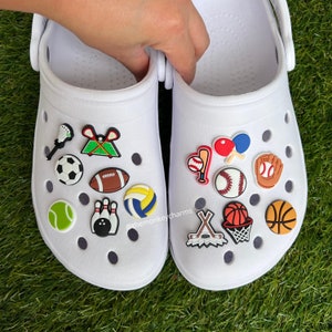 K&ouml;nnte beinhalten: Wei&szlig;e Clogs, verziert mit Sport-Themen-Anh&auml;ngern. Zu den Anh&auml;ngern geh&ouml;ren ein Fu&szlig;ball, Baseball, Basketball, Tennisball, Volleyball, Kegel und Hockeyschl&auml;ger. Die Clogs befinden sich auf einer gr&uuml;nen Oberfl&auml;che.