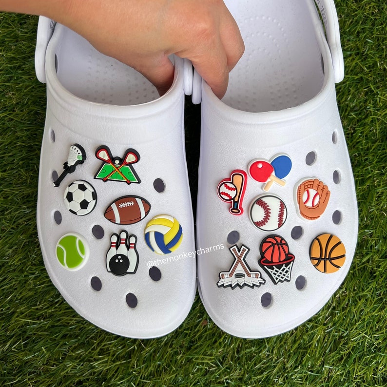 K&ouml;nnte beinhalten: Wei&szlig;e Clogs, verziert mit Sport-Themen-Anh&auml;ngern. Zu den Anh&auml;ngern geh&ouml;ren ein Fu&szlig;ball, Baseball, Basketball, Tennisball, Volleyball, Kegel und Hockeyschl&auml;ger. Die Clogs befinden sich auf einer gr&uuml;nen Oberfl&auml;che.