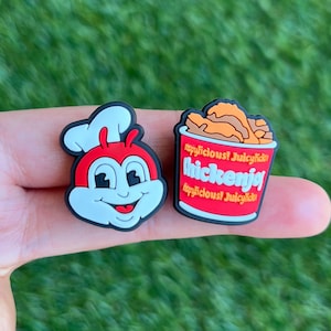 May include: Two colorful shoe charms. One is a smiling cartoon character in a chef's hat, with a red body and white face. The other is a red bucket of fried chicken with the words "chickenjoy" and "crispylicious" on it.