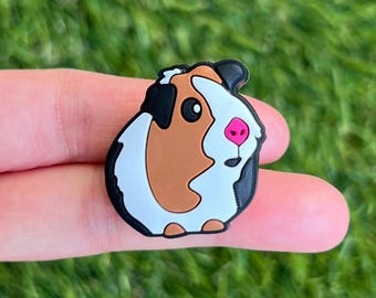 May include: A colorful guinea pig charm with a black outline, white, brown, and black fur, and a pink nose. The charm is a cute, cartoon-style design, perfect for animal lovers.