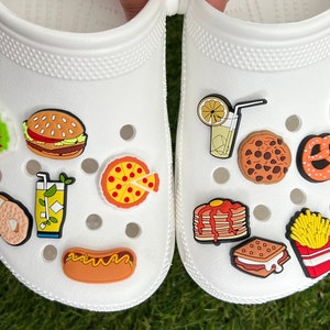 May include: White clogs decorated with colorful food-themed charms. The charms include a taco, burger, pizza slice, hot dog, fries, cookie, pretzel, and more. The clogs are on a green surface.