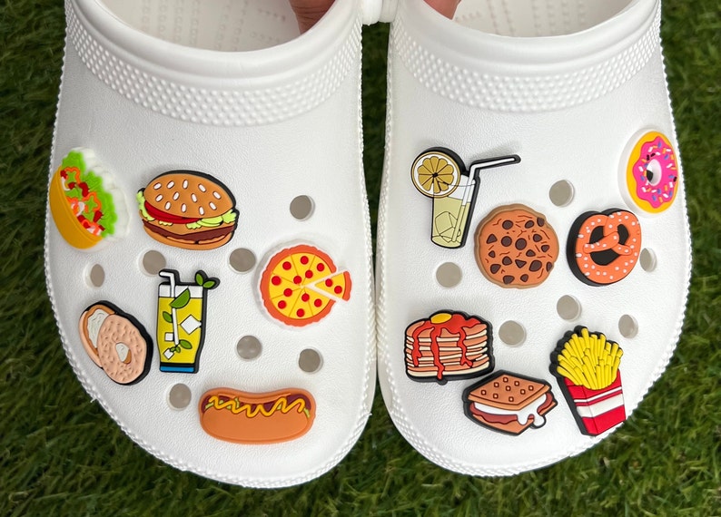 May include: White clogs decorated with colorful food-themed charms. The charms include a taco, burger, pizza slice, hot dog, fries, cookie, pretzel, and more. The clogs are on a green surface.