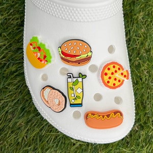 May include: White clog shoe decorated with colorful food-themed charms. Charms include a taco, burger, pizza slice, bagel, lemonade, and hot dog. The charms are in bright colors and add a playful touch to the shoe.