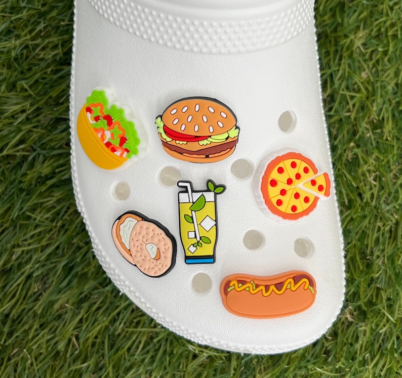 May include: White clog shoe decorated with colorful food-themed charms. Charms include a taco, burger, pizza slice, bagel, lemonade, and hot dog. The charms are in bright colors and add a playful touch to the shoe.