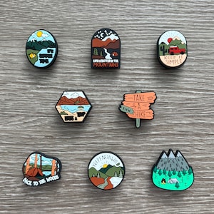 May include: A collection of camping-themed shoe charms. Designs include mountains, tents, and phrases like "It's Summer Time," "Life is Better in the Mountains," and "Keep it Simple." The charms feature various colors and shapes.