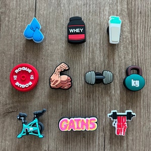 May include: A collection of colorful fitness-themed charms. Includes a red weight plate, a black and red protein powder container, a dumbbell, a kettlebell, a flexed arm, a water bottle, a stationary bike, and charms with the words "GAINS" and "NO DAYS OFF".