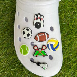 K&ouml;nnte beinhalten: Wei&szlig;er Clog-Schuh mit Sport-Themen-Anh&auml;ngern. Zu den Anh&auml;ngern geh&ouml;ren ein Fu&szlig;ball, Bowlingkegel, Tennisball, Volleyball, Lacrosse-Schl&auml;ger, ein Lacrosse-Schl&auml;ger und ein Fu&szlig;ball.