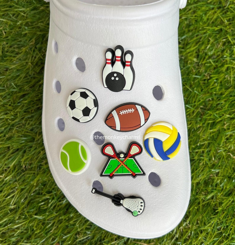 K&ouml;nnte beinhalten: Wei&szlig;er Clog-Schuh mit Sport-Themen-Anh&auml;ngern. Zu den Anh&auml;ngern geh&ouml;ren ein Fu&szlig;ball, Bowlingkegel, Tennisball, Volleyball, Lacrosse-Schl&auml;ger, ein Lacrosse-Schl&auml;ger und ein Fu&szlig;ball.