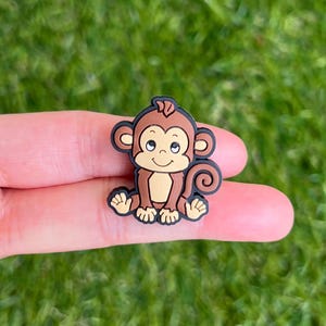 May include: A small, cartoon monkey charm in shades of brown, tan, and black. The monkey has a smiling face and is sitting upright. The charm is likely made of plastic or resin and is designed to be attached to a shoe or other item.