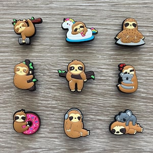 May include: Nine colorful sloth-themed charms in various poses. One sloth hangs from a branch, another floats on a unicorn floatie, and others are depicted with a koala, donut, and headphones. The charms are brown, tan, and black.
