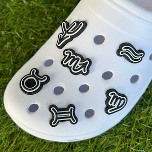 May include: White shoe with several black and white zodiac sign charms. The charms are in the shape of the zodiac symbols. The shoe is on a bed of green grass.