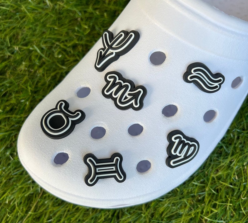 May include: White shoe with several black and white zodiac sign charms. The charms are in the shape of the zodiac symbols. The shoe is on a bed of green grass.