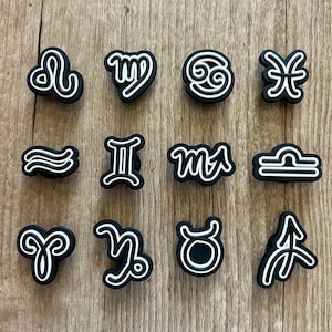 May include: A collection of black and white zodiac symbol charms. Each charm features a different astrological sign, outlined in white against a black background. The charms are arranged on a light brown wooden surface.