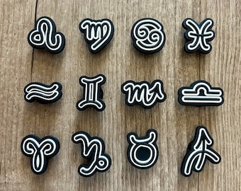 May include: A collection of black and white zodiac symbol charms. Each charm features a different astrological sign, outlined in white against a black background. The charms are arranged on a light brown wooden surface.