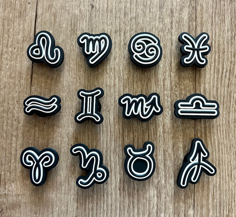 May include: A collection of black and white zodiac symbol charms. Each charm features a different astrological sign, outlined in white against a black background. The charms are arranged on a light brown wooden surface.