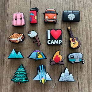 May include: A selection of camping-themed charms in various shapes and colours. Includes a pink backpack, a red thermos, an orange van, a black camera, a s'more, a marshmallow on a stick, a sign that says "I ❤️ CAMP", a guitar, mountains, a tent, a campfire, a camper, a pine tree, and skis.
