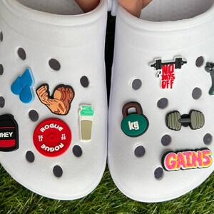May include: White Crocs shoes decorated with various fitness-themed charms. Charms include a weight plate, a protein shaker, a dumbbell, a kettlebell, and the word "GAINS". Other charms depict a flexing arm, water droplets, and a stationary bike.