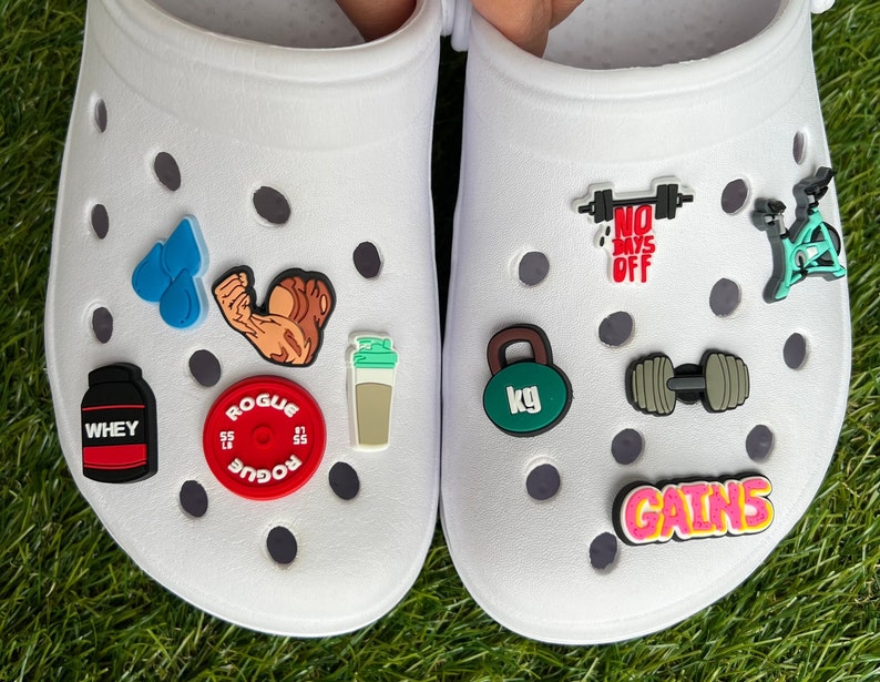 May include: White Crocs shoes decorated with various fitness-themed charms. Charms include a weight plate, a protein shaker, a dumbbell, a kettlebell, and the word "GAINS". Other charms depict a flexing arm, water droplets, and a stationary bike.