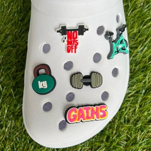 May include: White shoe with several fitness-themed charms. Charms include a green kettlebell with "kg" text, a dumbbell, a stationary bike, a barbell with "NO DAYS OFF" text, and a "GAINS" text charm.