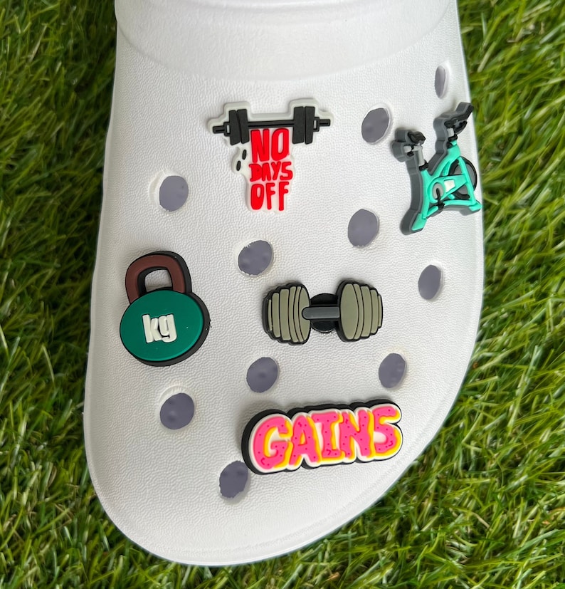 May include: White shoe with several fitness-themed charms. Charms include a green kettlebell with "kg" text, a dumbbell, a stationary bike, a barbell with "NO DAYS OFF" text, and a "GAINS" text charm.