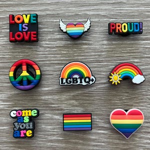 May include: A collection of colorful shoe charms with LGBTQ+ pride themes. Designs include a rainbow heart with wings, a rainbow peace sign, a rainbow with a sun and cloud, and charms with the phrases "LOVE IS LOVE", "PROUD!", and "COME AS YOU ARE".