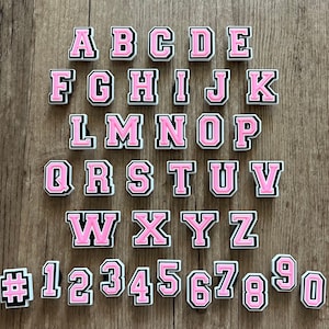 May include: A collection of pink and white alphabet and number charms. Each charm has a pink center with a white border and a black outline. The letters and numbers are arranged on a wooden surface.