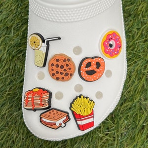 May include: White shoe with various food-themed charms. Charms include a lemon drink, chocolate chip cookie, pretzel, donut, stack of pancakes, s'more, and a box of french fries. The charms are colorful and add a playful touch.