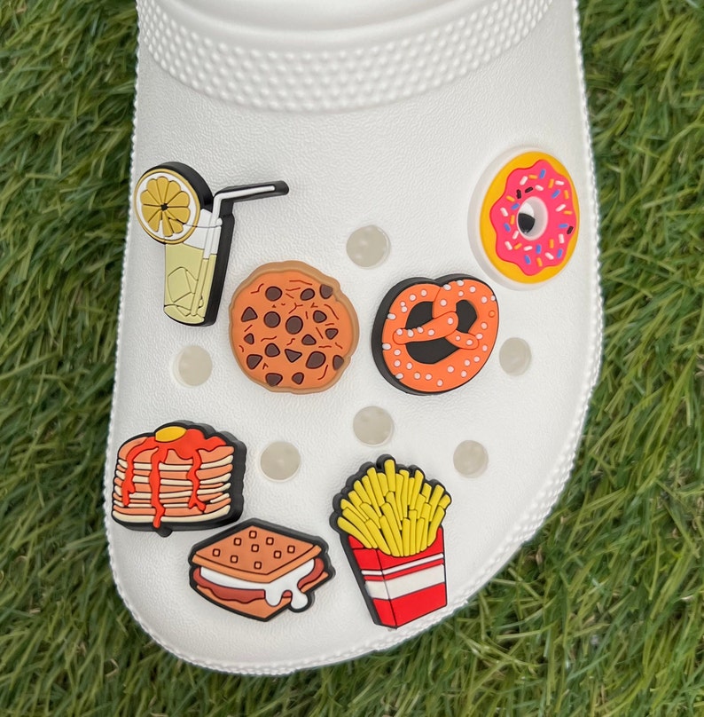 May include: White shoe with various food-themed charms. Charms include a lemon drink, chocolate chip cookie, pretzel, donut, stack of pancakes, s'more, and a box of french fries. The charms are colorful and add a playful touch.