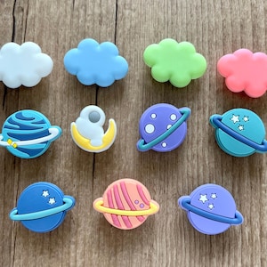 May include: A collection of colorful shoe charms featuring space and weather themes. The charms include white, blue, green, and pink cloud shapes, along with planets in blue, purple, and pink, and an astronaut on a crescent moon. These charms are designed to decorate shoes.