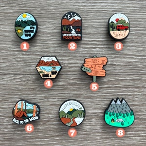 May include: A collection of eight camping-themed shoe charms. Each charm features a different design, including mountains, tents, and phrases like "Keep It Simple" and "Life is Better in the Mountains." The charms are in various shapes and colors.